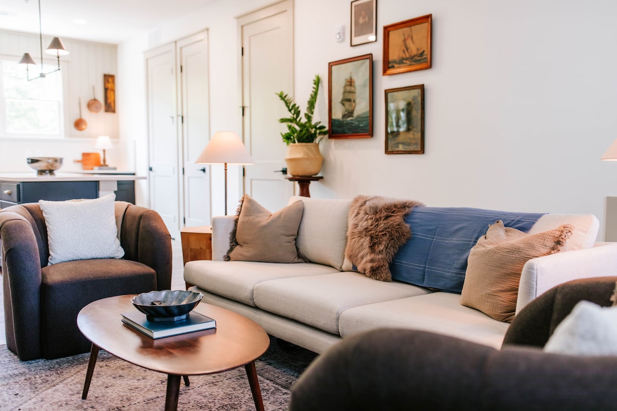 A cozy lounge area is presented with a light-colored sofa adorned with an assortment of pillows. Flanking the sofa are two round dark armchairs. Warm lighting is provided by two elegant lamps, while framed artwork hangs on the wall, enhancing the inviting ambiance.