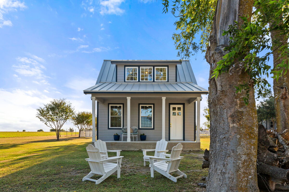 A charming tiny home is showcased, featuring a metal roof and welcoming front porch. Four white Adirondack chairs are arranged around a grassy area, offering a view of the expansive countryside. Trees frame the scene, contributing to the serene environment.