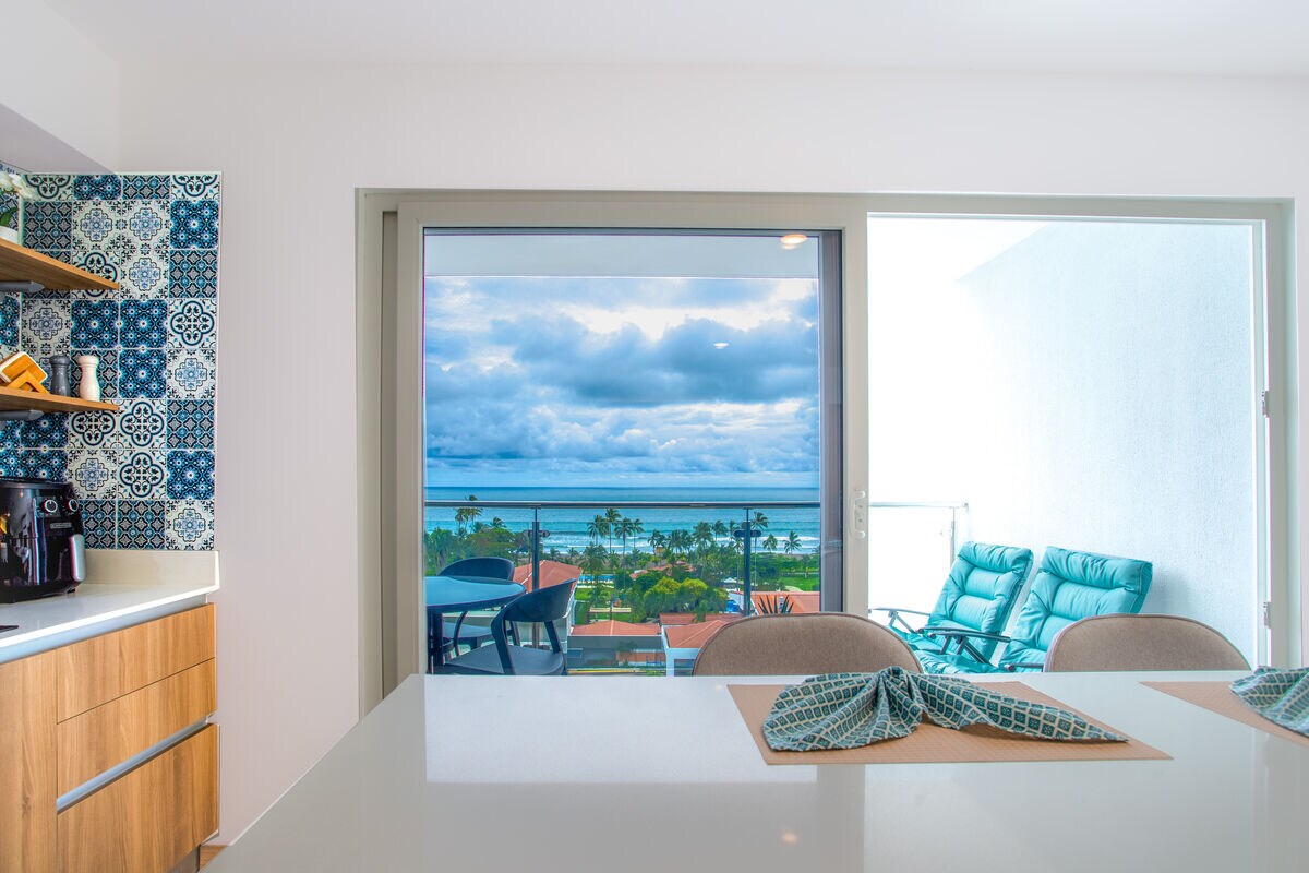 A view of the kitchen area features modern cabinetry and a breakfast bar. A large sliding glass door opens to a private balcony, showcasing ocean views and a seating area. The blue tones of the decor complement the coastal scenery seen beyond.