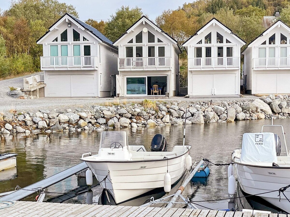 A row of three holiday homes is seen along the marina, each featuring large windows and a balcony. A small motorboat is docked nearby, with a floating dock extending into the calm water. Lush greenery is visible on the surrounding hillside.