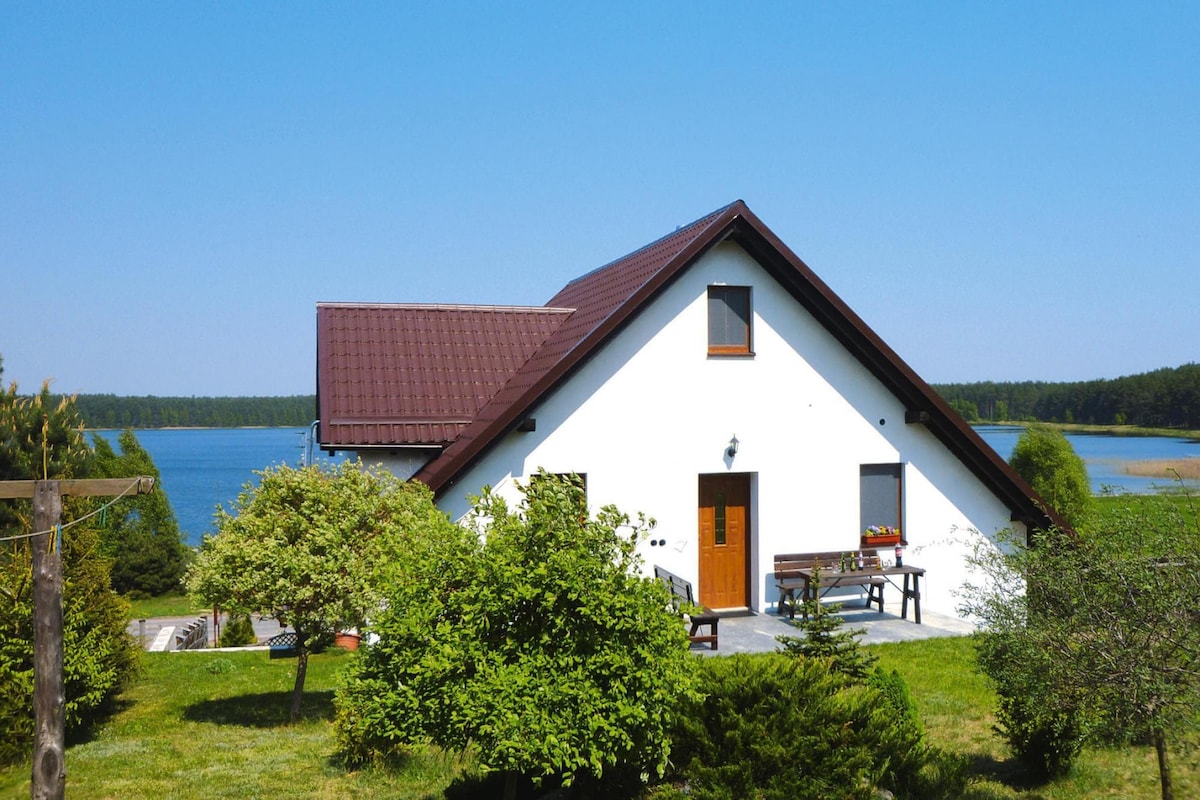 A white house with a sloped roof is situated near a tranquil body of water. Lush greenery surrounds the property, with trees and shrubs enhancing the natural setting. A wooden table and chairs are positioned outside, offering a space for outdoor relaxation.
