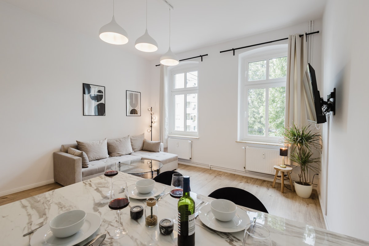 A light-filled living area features a comfortable sofa positioned near large windows, offering views of greenery outside. A stylish dining table is set for a meal, showcasing plates, glasses, and a wine bottle. Minimalist decor is present, with wall art adding subtle interest.