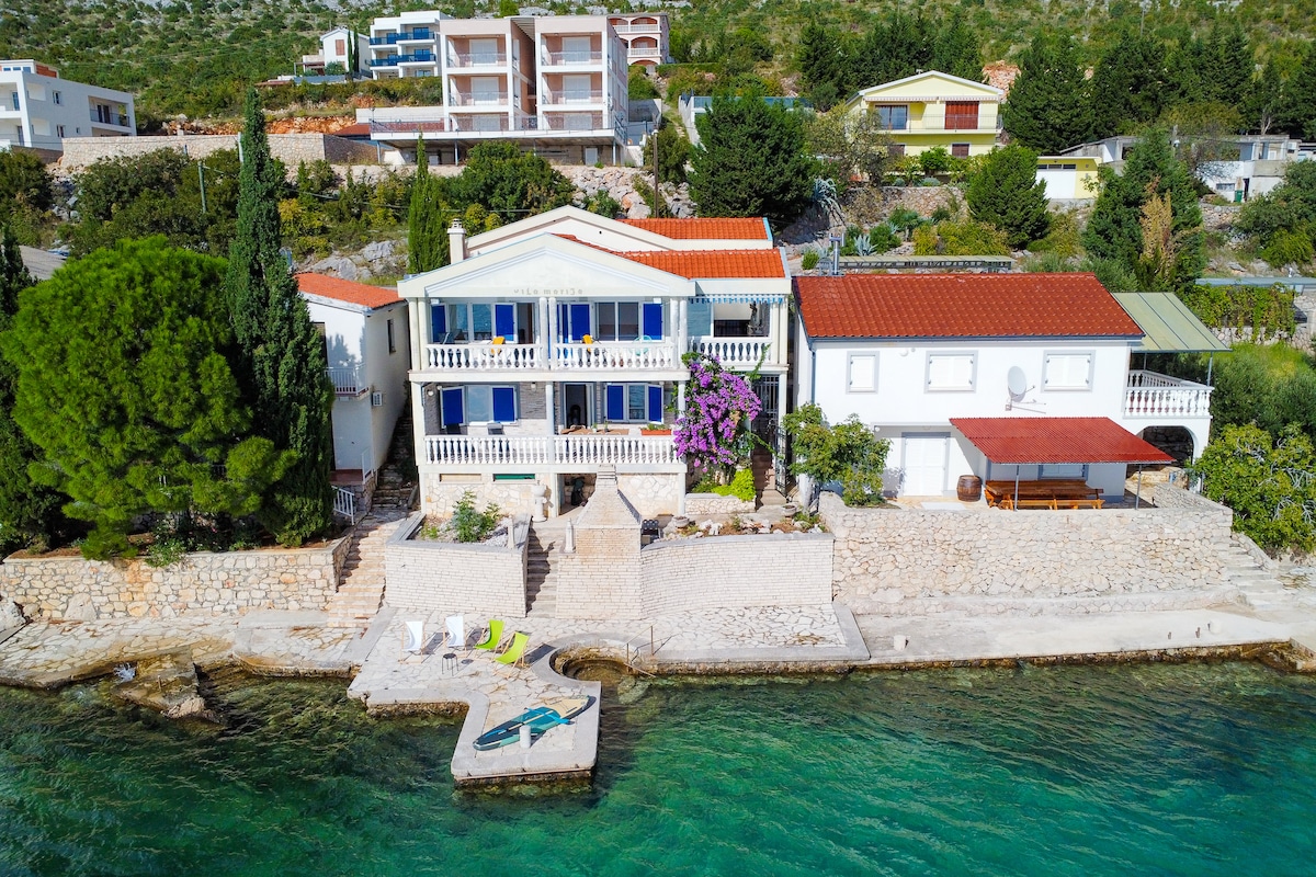 The villa is situated directly along the shoreline, featuring balconies adorned with vibrant flowers. The outdoor area includes a patio and sun loungers by the water, creating an inviting space for relaxation. Nearby residential buildings are visible in a scenic coastal setting.
