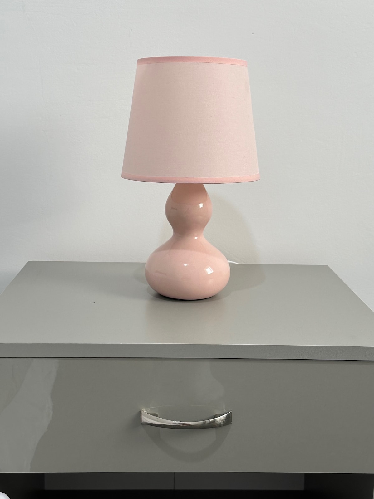 A soft pink lamp with a curved base and matching lampshade is placed on a modern gray nightstand. The smooth surface of the nightstand reflects light, enhancing the overall design of the space.