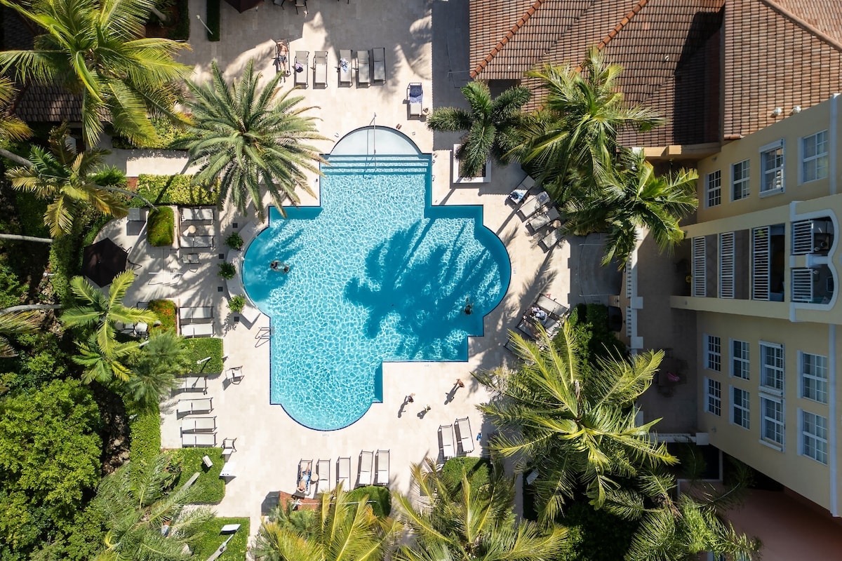 An aerial view captures a resort-style pool surrounded by lush palm trees and lounge chairs. The pool's unique shape is highlighted, with shadows cast over the water. Several guests can be seen enjoying the space, creating a serene environment.