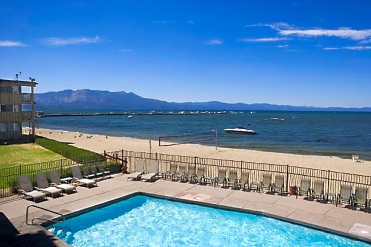 A clear view of the pristine beach and Lake Tahoe can be seen from the property. A swimming pool sits in the foreground, surrounded by loungers and a volleyball court. The distant mountains and a boat on the water add to the serene landscape.
