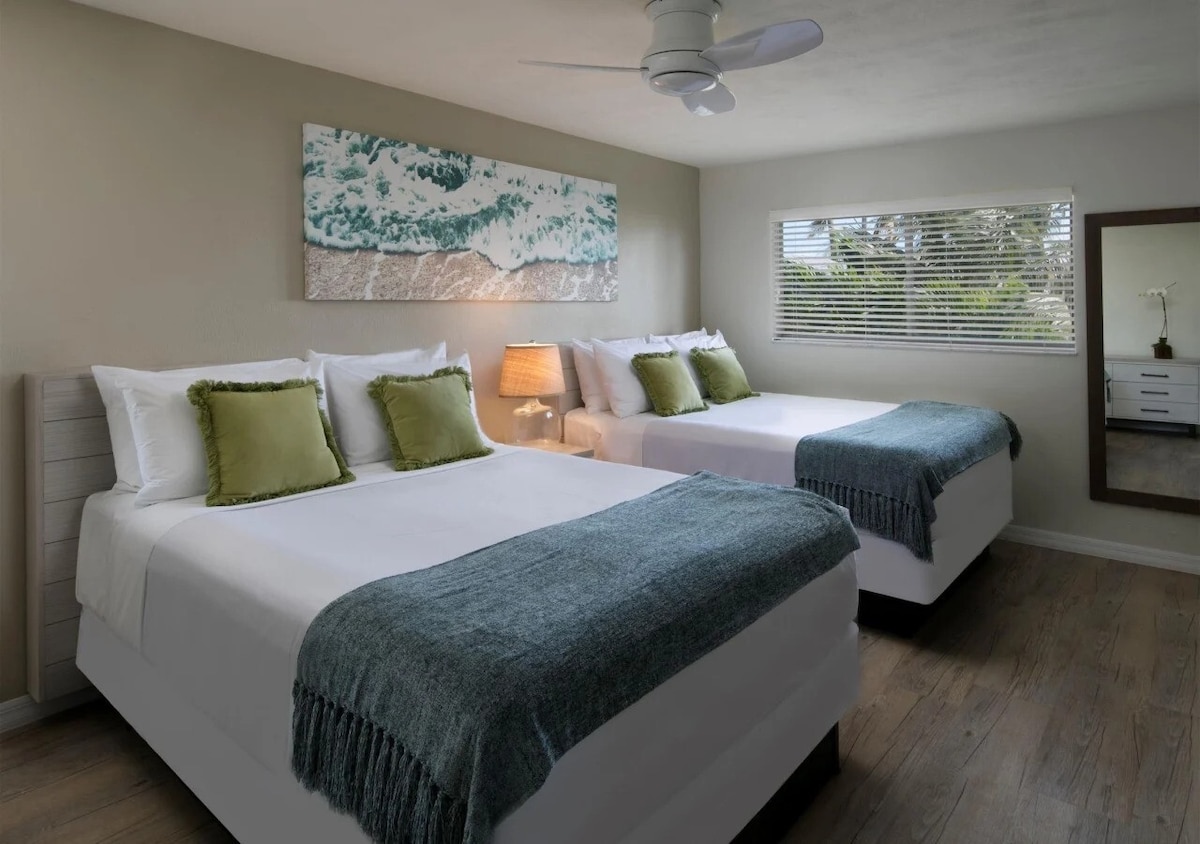 A well-lit bedroom features two spacious queen beds dressed with soft white linens and green accent pillows. A textured throw blanket drapes over each bed. A small bedside lamp provides warm lighting, while a window offers natural light and a view of garden greenery outside.