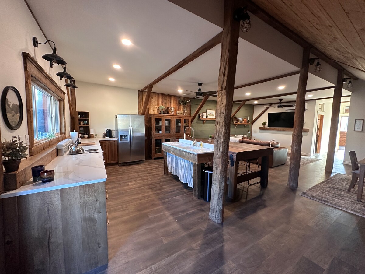The spacious kitchen area features wooden cabinetry and a stainless-steel refrigerator. Natural light streams through the window above the sink, highlighting the rustic wood beams and open layout of the living space. A large dining table is positioned nearby, complemented by comfortable seating.