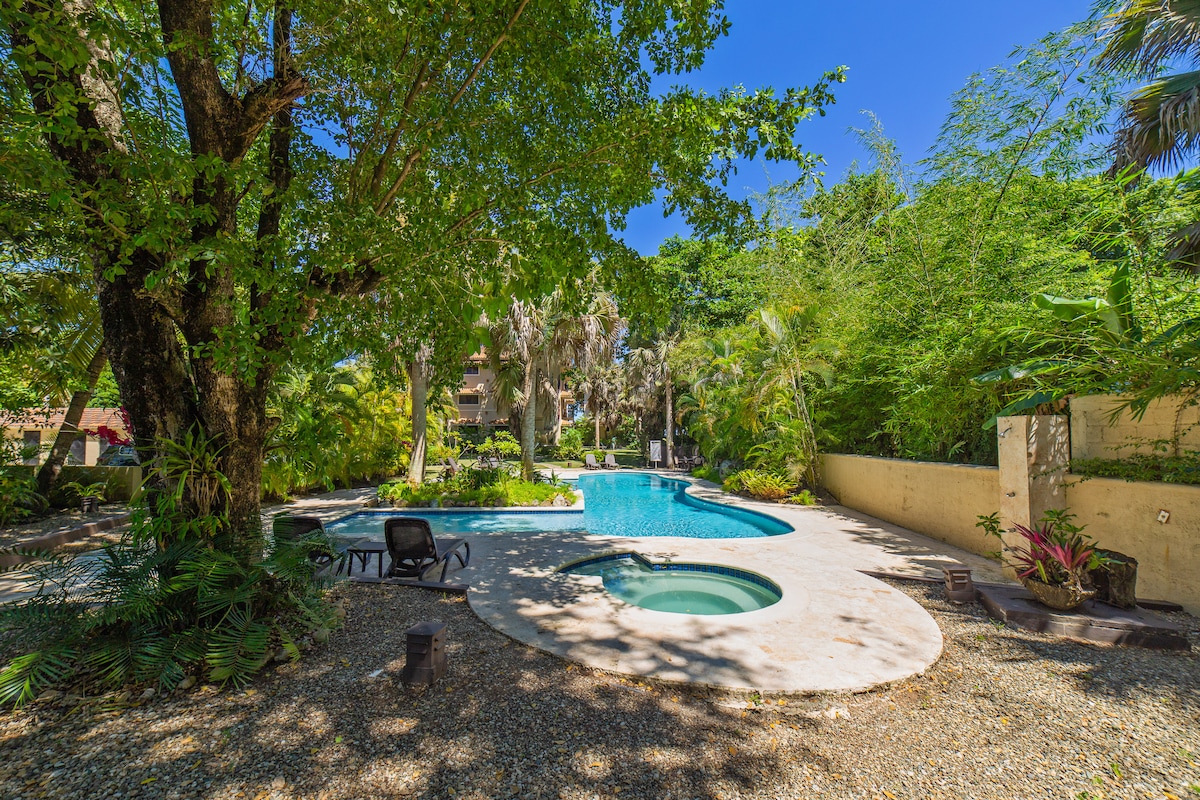 A spacious swimming pool is surrounded by lush tropical greenery and palm trees, creating a tranquil setting. The pool features a circular spa, and a shaded area with lounge chairs is positioned conveniently nearby, inviting relaxation in a serene outdoor environment.