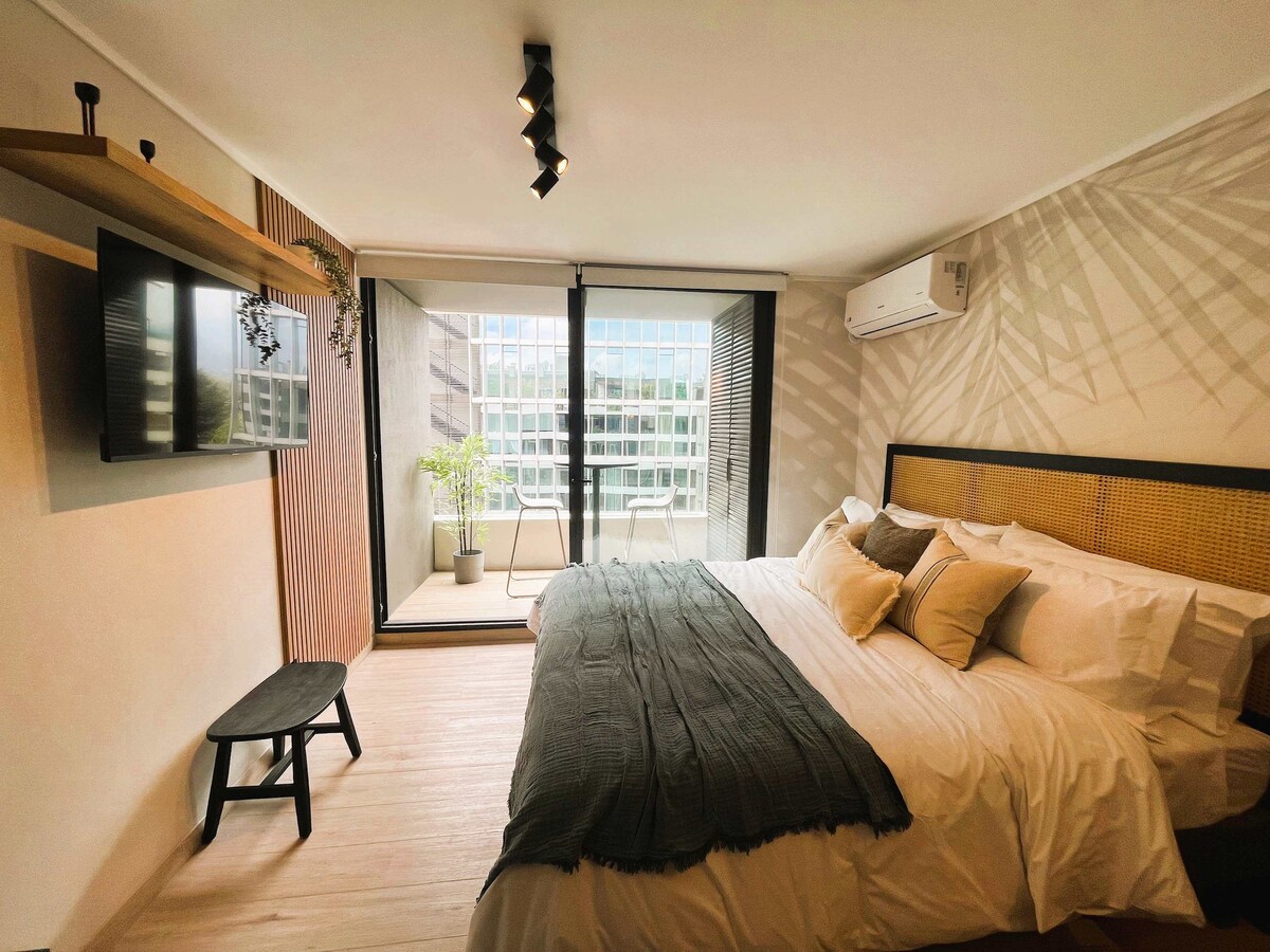 A well-lit sleeping area is highlighted by a spacious bed with soft white linens and a dark textured throw. Natural light floods in through glass doors that open to a private terrace, accompanied by a small potted plant and a minimalist bench nearby.