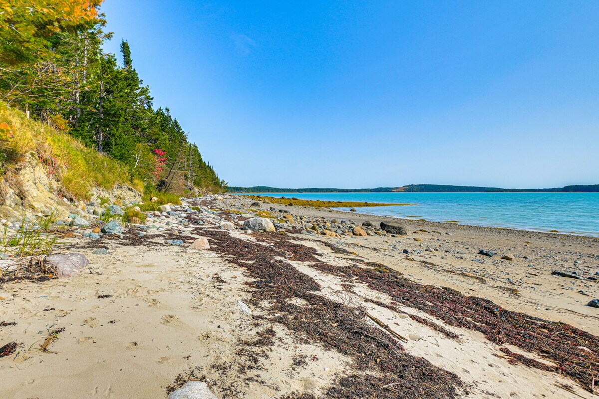 A peaceful beach scene is presented, marked by a mix of sand and pebbles along the shoreline. Gentle waves meet the shore beneath a clear blue sky, with lush greenery and rocky outcrops visible in the background. The scenery reflects a tranquil coastal environment.
