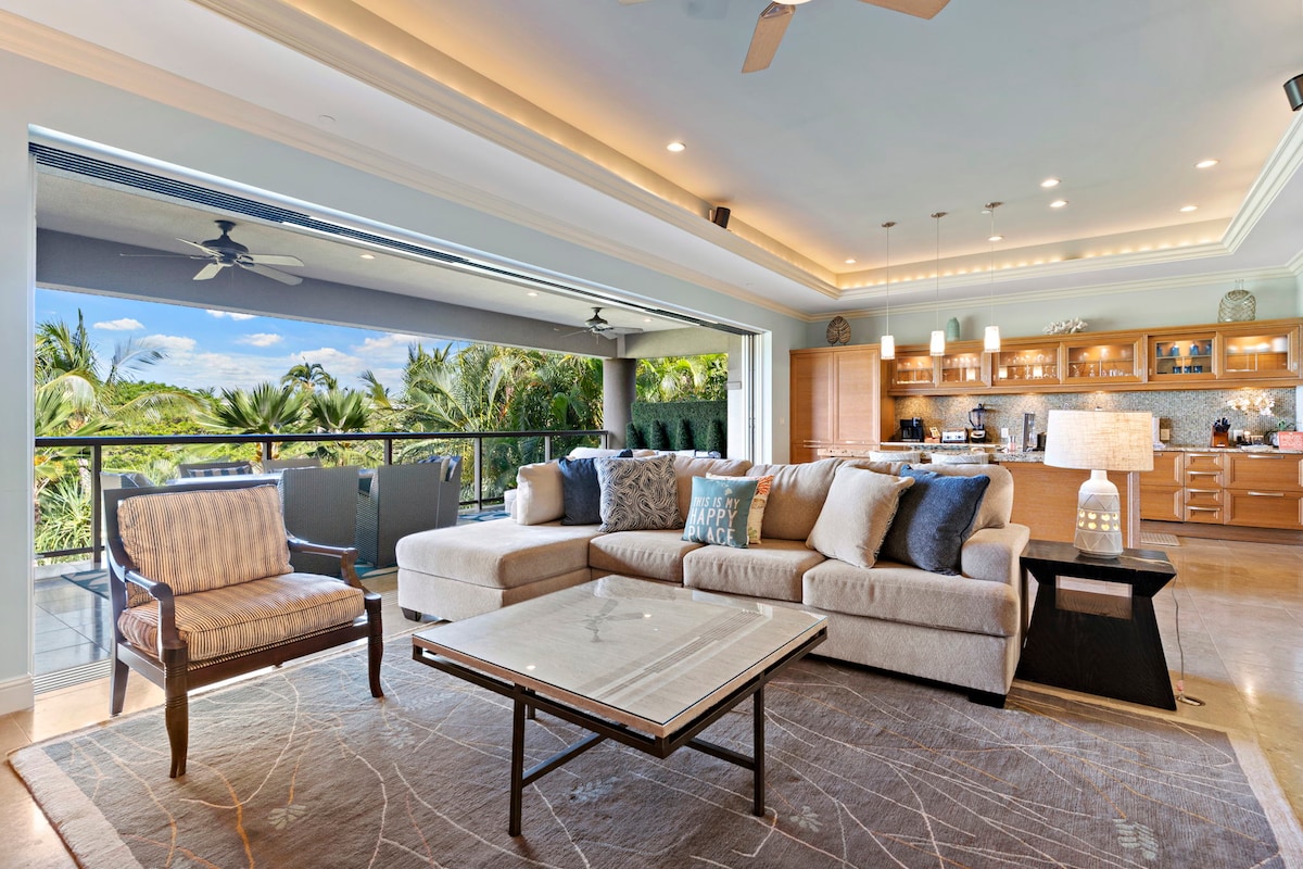 The open living area features a spacious sectional sofa, a glass coffee table, and a decorative rug. Large sliding doors lead to an outdoor space framed by tropical greenery, allowing natural light to fill the room. The kitchen area with wooden cabinetry and modern appliances is visible in the background.