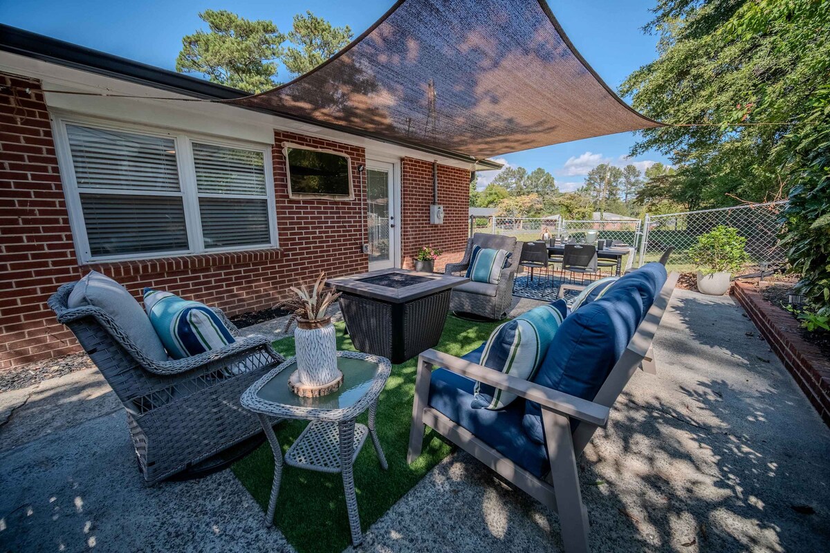 An outdoor patio area features comfortable seating arranged around a central fire table. A shaded space is created by a canopy overhead. Lush greenery is visible in the background, along with a fenced yard that enhances privacy.