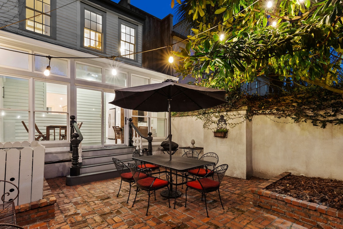 A private outdoor courtyard is illuminated by charming string lights, featuring a dining table for six beneath a large umbrella. Surrounding brick flooring adds warmth, complemented by a magnolia tree that enhances the outdoor ambiance.