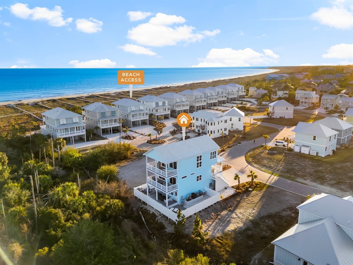 An aerial view showcases a coastal retreat surrounded by vibrant greenery and other homes. The serene Gulf waters are visible in the distance. Beach access is highlighted, emphasizing the home's proximity to the shoreline. The retreat features a light blue exterior, blending with the beach scene.