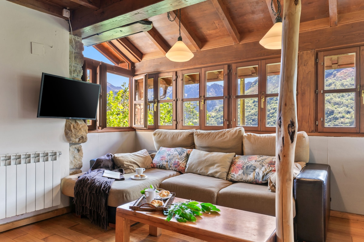 A comfortable living room is highlighted by a large sectional sofa adorned with floral cushions. Several wooden windows provide a view of the mountains. A coffee table sits in the center, displaying pastries and foliage, while a flat-screen TV is mounted on the wall.