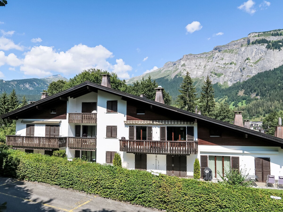 The exterior of the villa features a three-story structure with a combination of wood and white facades. Several balconies are visible on the upper floors, surrounded by greenery and with mountain views in the background under a clear blue sky.