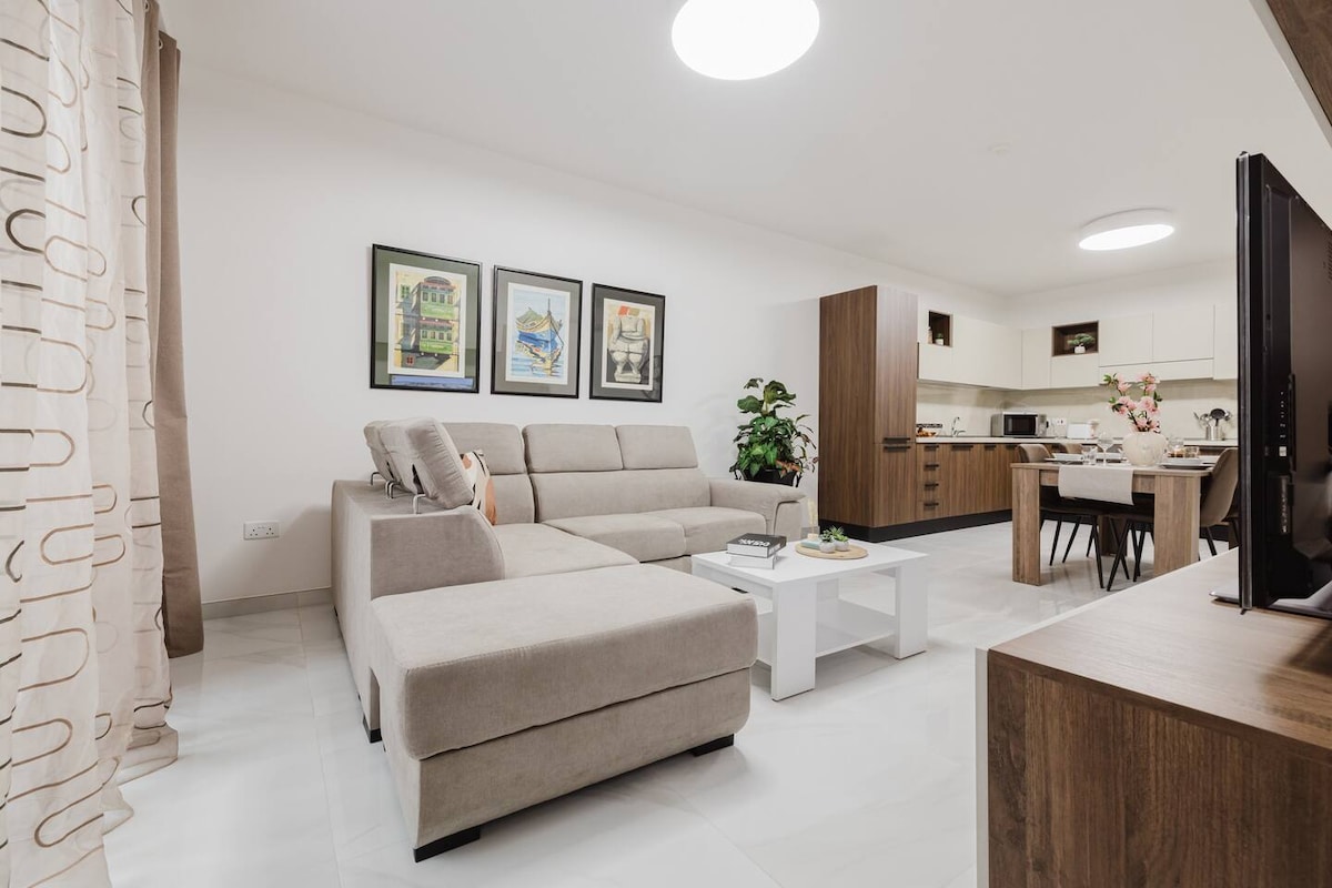 A modern living area features a comfortable beige sectional sofa and a minimalist coffee table atop tiled flooring. Framed artwork adorns the wall, adding character. A dining table and fully equipped kitchen are visible in the background, showcasing an inviting open layout.
