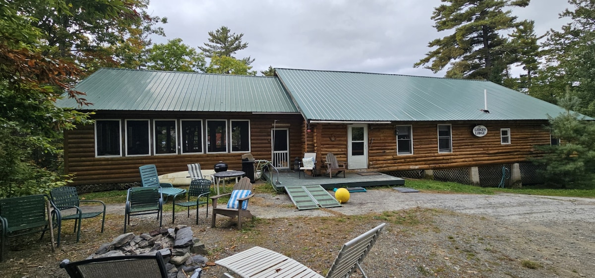 A spacious log cabin exterior showcases a green metal roof and wooden siding surrounded by trees. A deck leads to the entrance, while several outdoor chairs and a fire pit are positioned in the yard, inviting relaxation and gatherings.