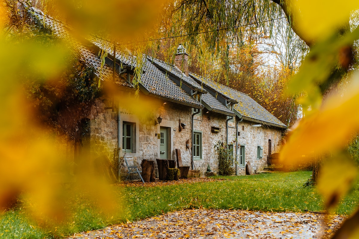 An ancient stone cottage is framed by colorful autumn leaves. The building features multiple windows and a textured roof, set amidst a lush green lawn. The entrance is adorned with rustic pots, enhancing the connection with nature surrounding the home.