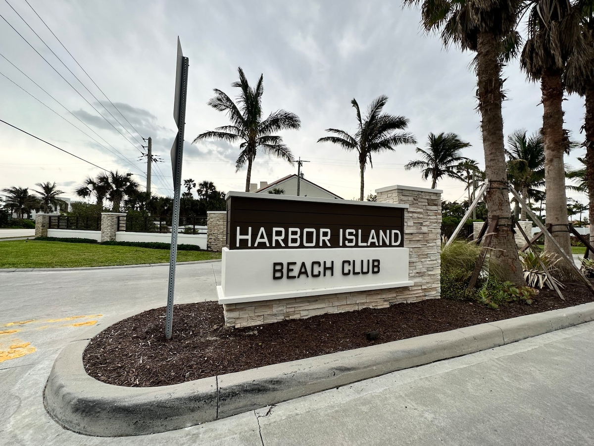 A welcoming entrance sign for Harbor Island Beach Club is highlighted in front of palm trees and well-maintained greenery. The sign features the name prominently displayed in bold letters, signaling the start of a serene beach community experience.