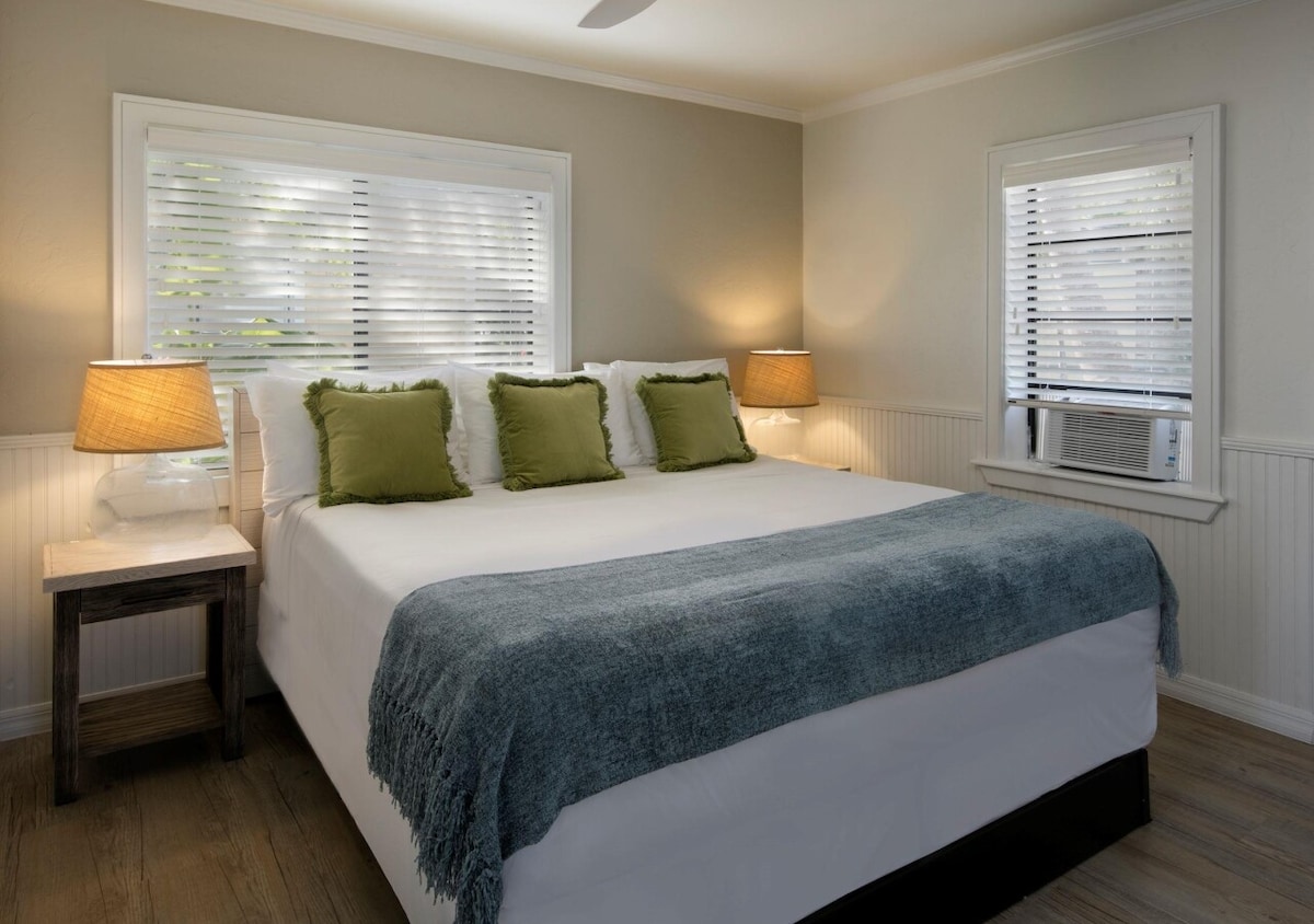 A spacious bedroom features a large bed topped with plush pillows and a cozy throw blanket. Two bedside lamps provide soft lighting, and large windows with blinds allow natural light to filter in. A wall-mounted air conditioning unit is visible near the window.
