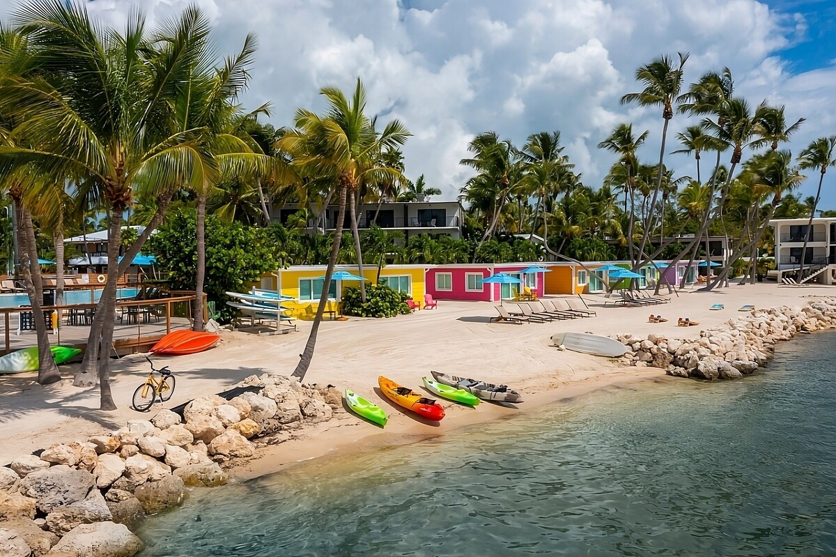 A sandy beachfront is lined with colorful cottages surrounded by palm trees. Several kayaks in vibrant colors rest on the shore, while a lone bicycle is parked nearby. The clear water reflects the sky, enhancing the tropical ambiance.