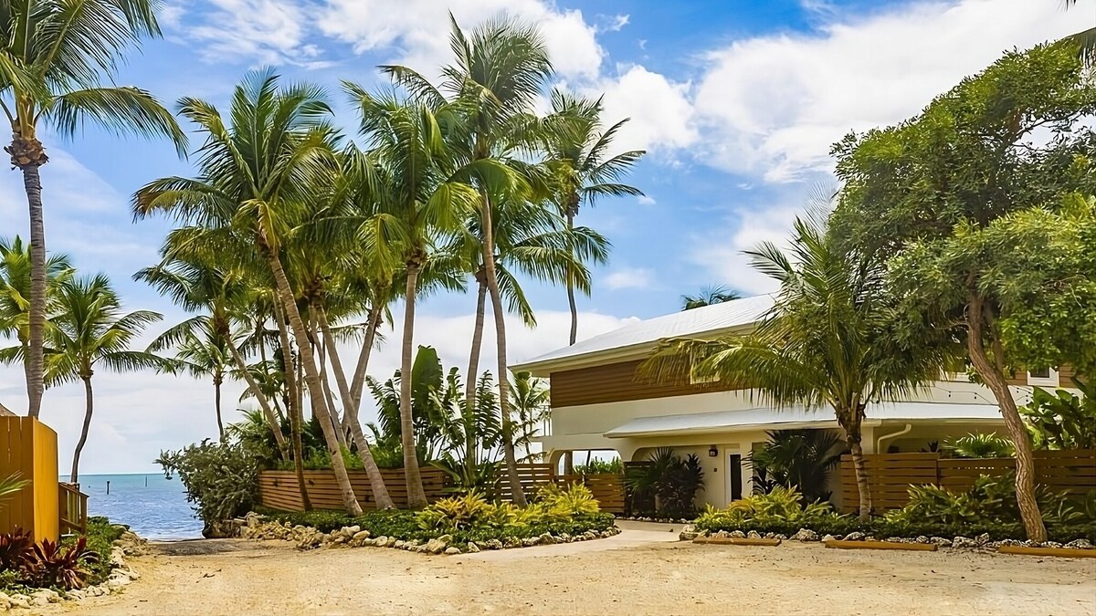 A tropical exterior view of the property is presented, surrounded by lush palm trees and vibrant landscaping. The main building features a modern design with a light-colored facade and a large porch area, set against a backdrop of clear blue skies and the nearby ocean.