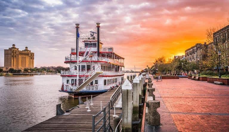 A historic riverboat is docked along a wooden pier, with vibrant colors of sunset reflecting on the water. A scenic pathway runs adjacent to the river, lined with trees and buildings, creating a tranquil riverside atmosphere.