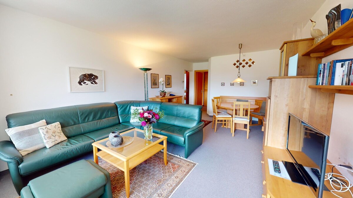 A spacious living-dining area features a green leather sectional sofa, a wooden coffee table, and a flat-screen TV. A dining table with chairs is visible in the background. Natural light illuminates the room, complemented by wall art and decorative shelving.