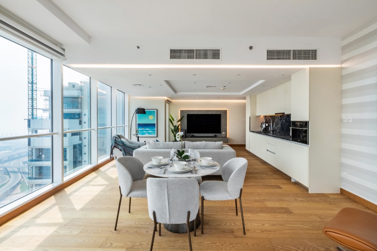A modern living area features large floor-to-ceiling windows, allowing natural light to illuminate the spacious room. A round dining table with four soft chairs is set for a meal. Designer decor is visible, along with a wall-mounted TV and a sleek kitchen area.
