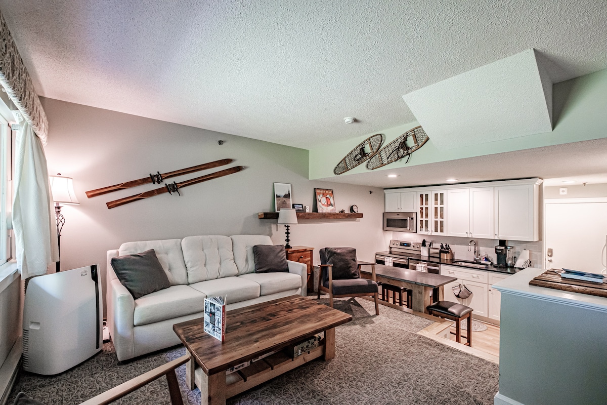 A cozy living area is highlighted by a comfortable couch and armchairs arranged around a wooden coffee table. Above, vintage skis are displayed on the wall. The modern kitchen features stainless appliances and light cabinetry, creating an open, inviting atmosphere.