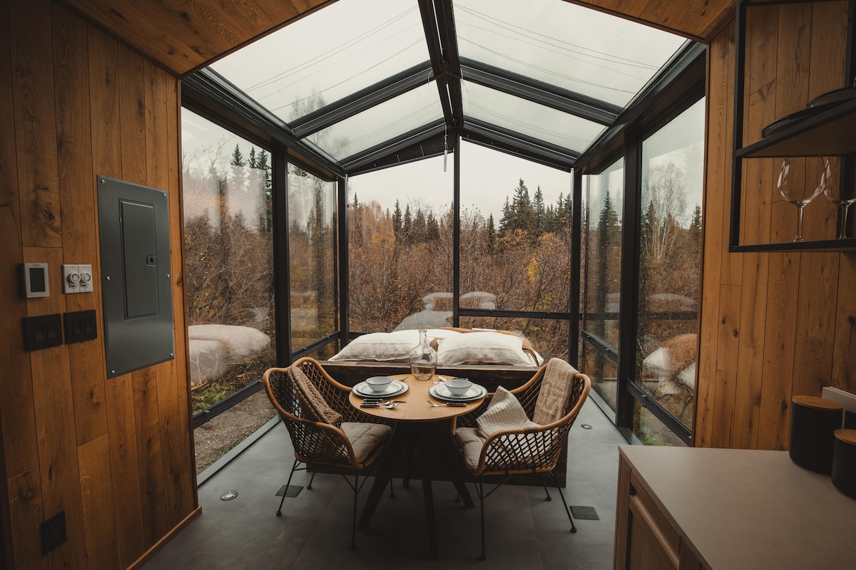 A cozy dining area features a round table set for two, surrounded by stylish rattan chairs. Large glass walls provide panoramic views of the surrounding nature, allowing natural light to enhance the wooden interior. Two comfortable beds are visible beyond the table, creating an inviting space.