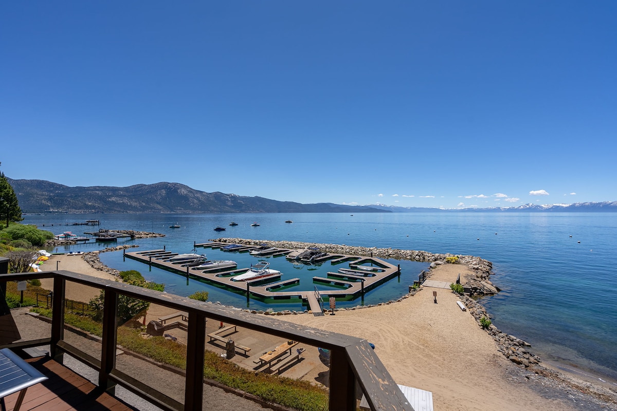 A panoramic view of Lake Tahoe is visible from a deck. The clear blue water extends towards a marina, where several boats are docked. A sandy beach is situated nearby, framed by rocky edges and lush greenery, creating a serene lakeside atmosphere.