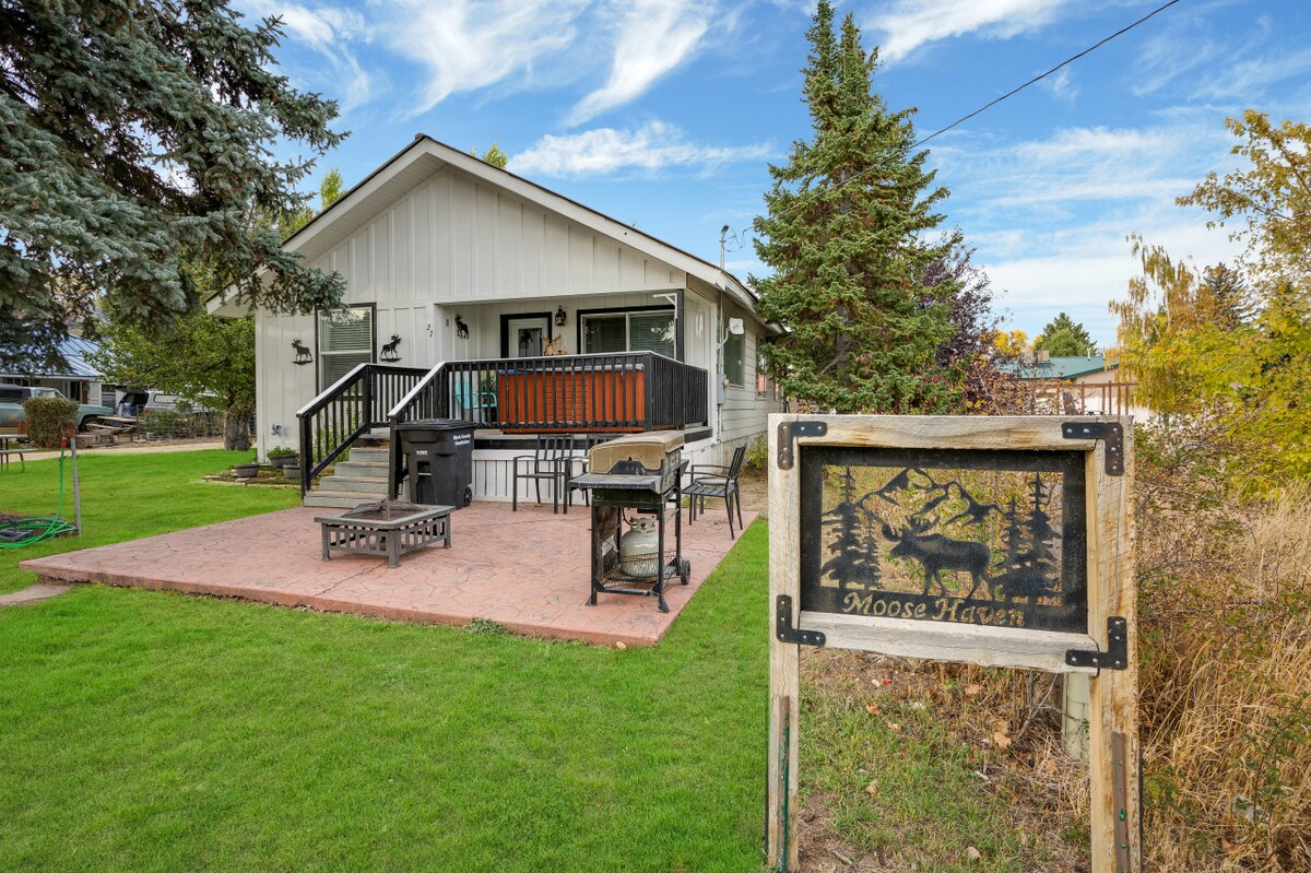 The exterior of a welcoming home is shown, featuring a well-maintained lawn and a patio area with outdoor seating. A wooden sign labeled 'Moose Haven' is positioned at the front, surrounded by a variety of trees and seasonal foliage.