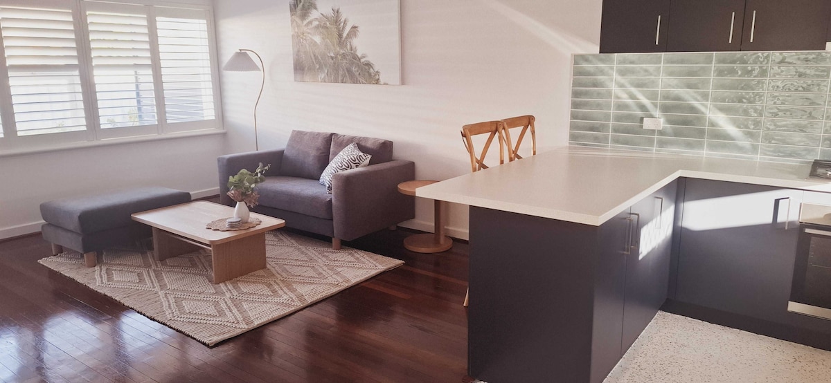 A spacious living area features a comfortable sofa and a light-colored coffee table atop a textured rug. Natural light filters through plantation shutters, highlighting the elegant kitchen area with dark cabinetry and a light countertop.