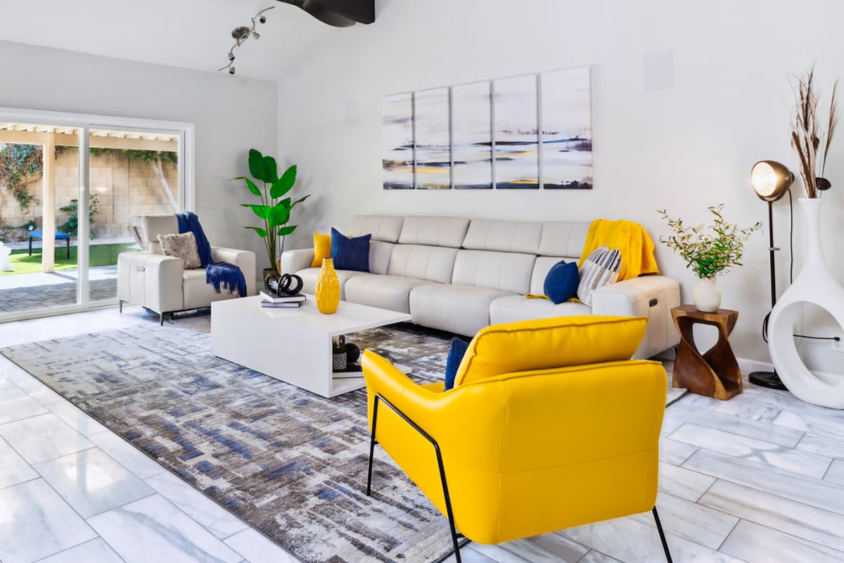 The spacious living area is furnished with a modern sectional sofa and a vibrant yellow accent chair. Light neutral walls and a colorful artwork piece enhance the space. Large sliding glass doors allow natural light and provide a view of the outdoor area.