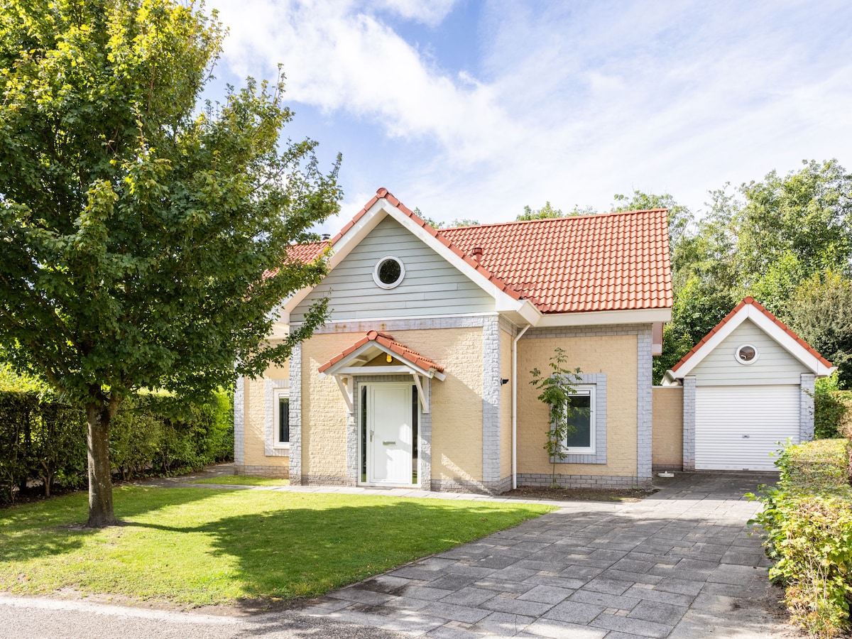 A single-story house with a light-colored façade and a red-tiled roof is framed by neatly trimmed hedges and trees. A spacious driveway leads to an attached garage on the right side. Clear blue skies create a bright setting for the home.