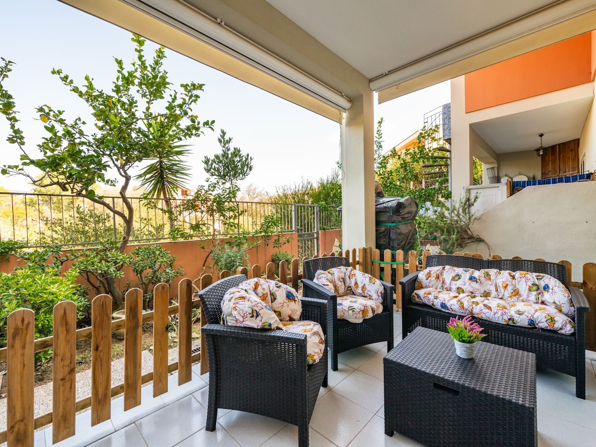 An outdoor seating area is furnished with a set of dark wicker chairs and a coffee table, adorned with floral cushions. The fenced small garden features various plants and trees, providing a natural backdrop. Sunlight streams in, creating a welcoming atmosphere.