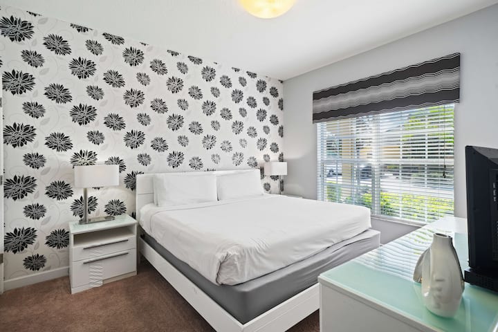 - Модерна спална соба со брачен кревет со свежа бела постелнина, врамена со задебелен црно-бел ѕид со цветен акцент - голем прозорец пропушта природна светлина, со шарена валанс која додава стилски допир - минималистичкиот декор создава свежа атмосфера