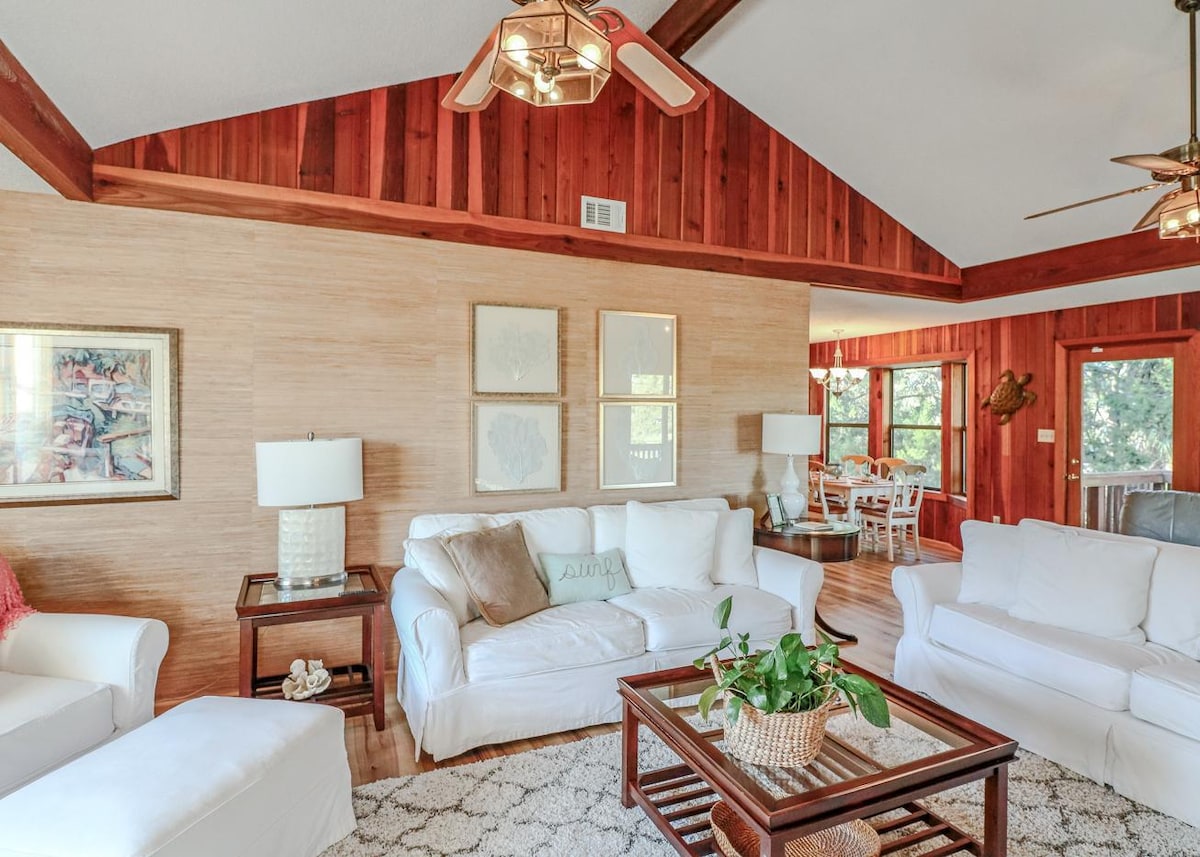 A spacious living room features a combination of white furniture and warm wooden accents. Two ceiling fans provide airflow, and a coffee table with a woven basket is centered on a light area rug. Natural light is welcomed through windows leading to the dining area.