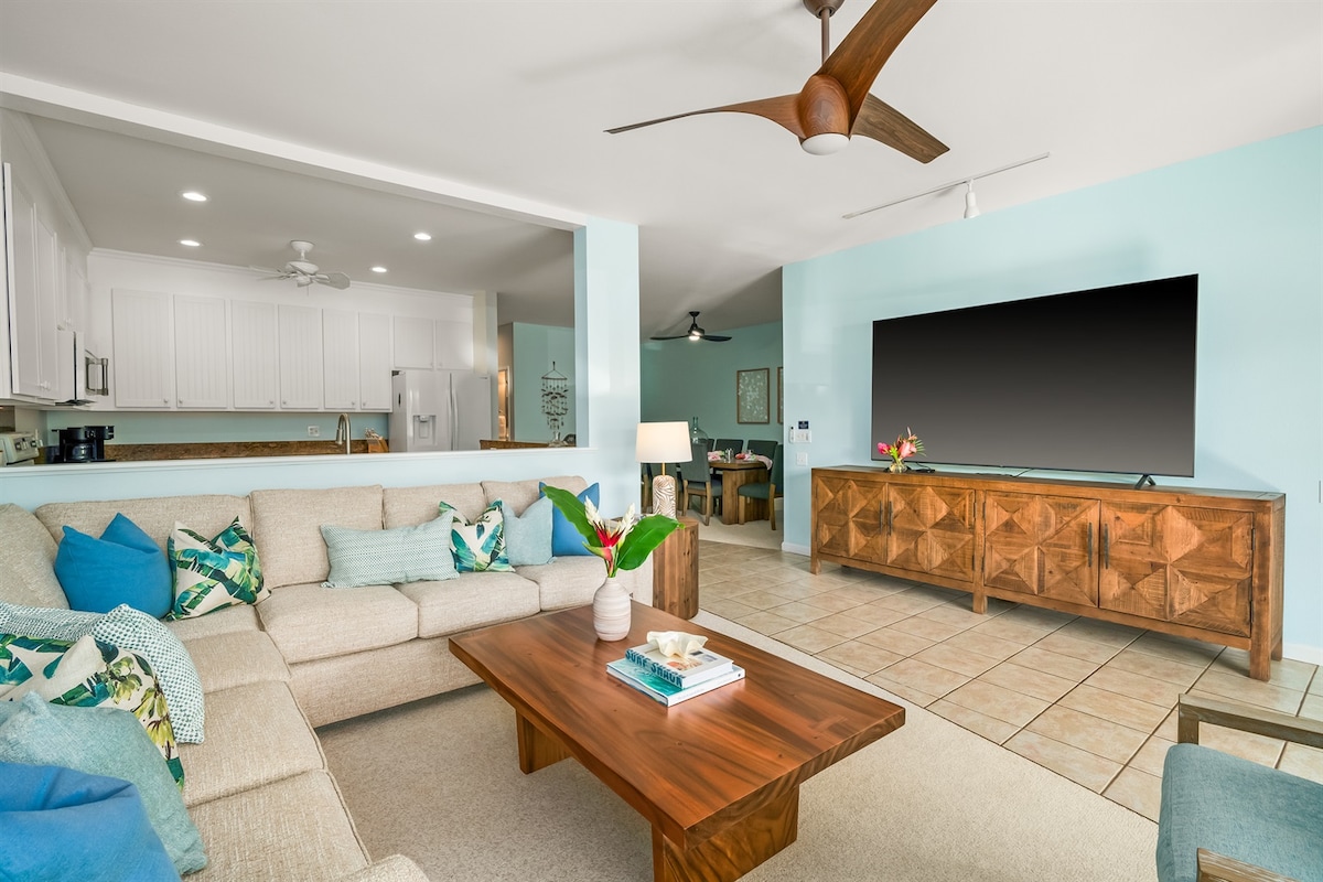 A spacious living area features a sectional sofa adorned with colorful cushions, complemented by a wooden coffee table. A large flat-screen TV is mounted on a wooden console, and a ceiling fan provides airflow. The open layout connects to the kitchen and dining areas.