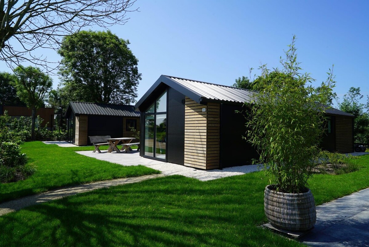 A modern chalet is surrounded by a lush green lawn, featuring a sleek design with a combination of dark and natural wood finishes. The spacious outdoor area includes a patio, as well as a picnic table, offering a comfortable setting for relaxation.