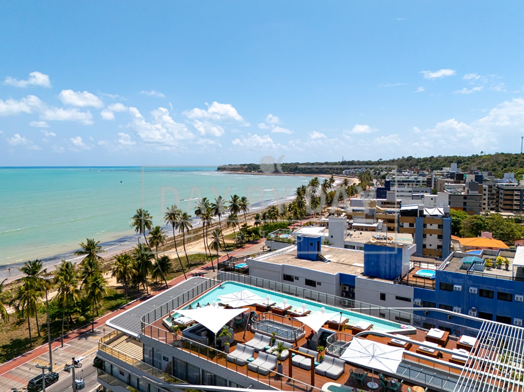 A panoramic view showcases a coastal area with a sandy beach lined by tall palm trees and urban buildings. The rooftop pool area with sun loungers is visible, providing a relaxing space with direct access to the scenic beachfront.