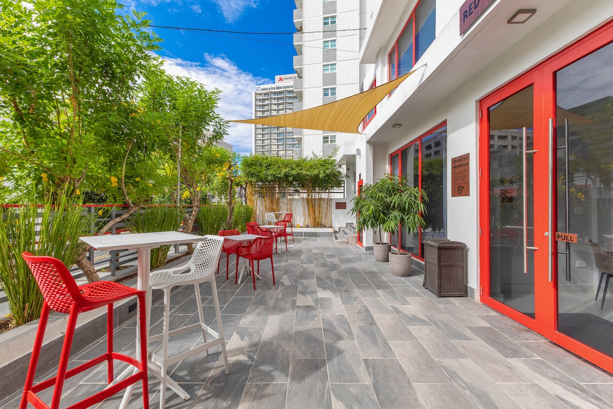 An outdoor seating area features a mix of red and white chairs and tables arranged on stone flooring. Lush greenery and potted plants create a refreshing ambiance, with a sunshade providing partial coverage. The exterior walls are adorned with large windows and bright red doors.