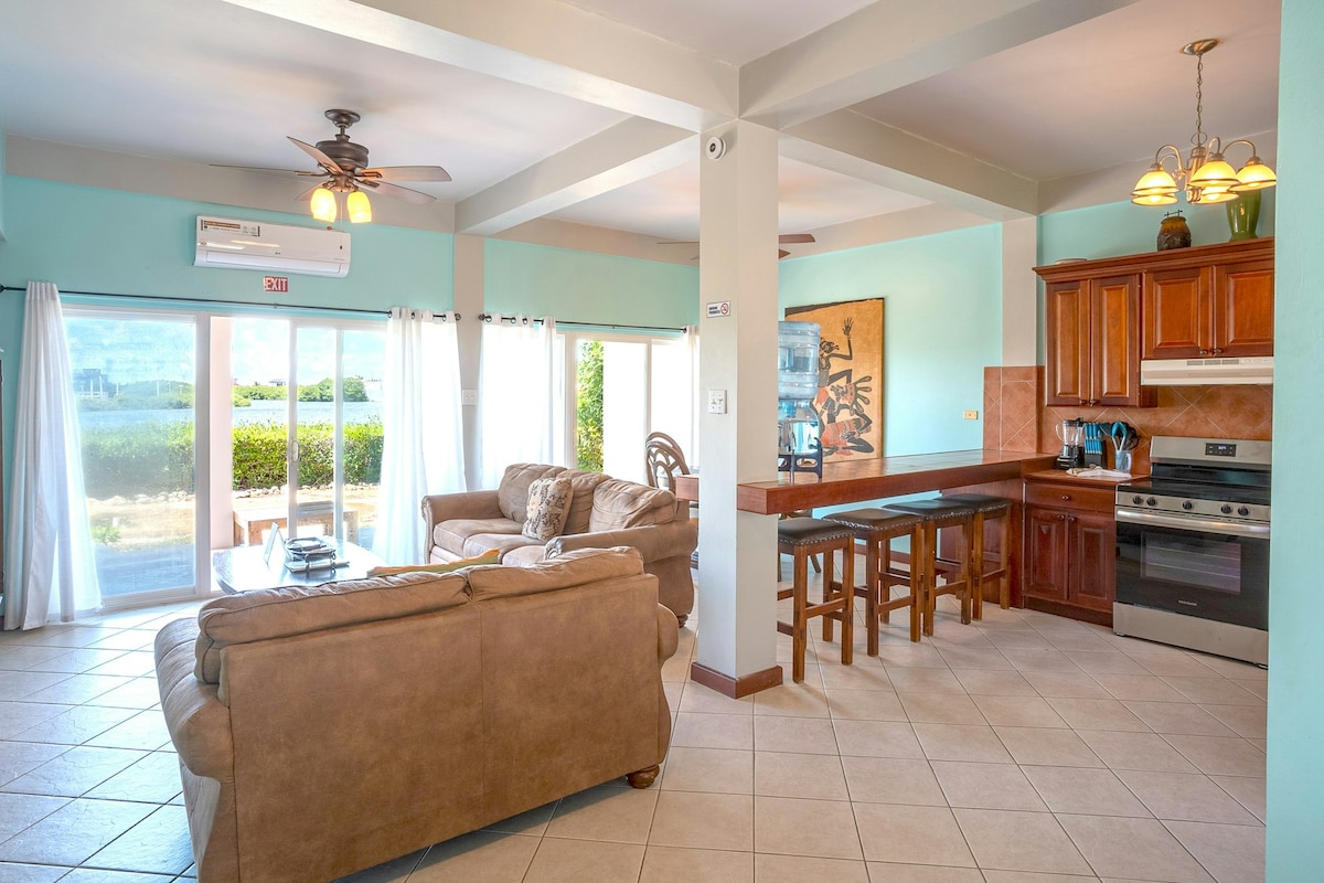 An open-plan living area is visible, featuring a comfortable sofa suite and a wooden dining table with three chairs. Natural light enters through large sliding glass doors, which lead to the outdoor space, highlighting the warm tones of the cabinetry and decor in the kitchen area.