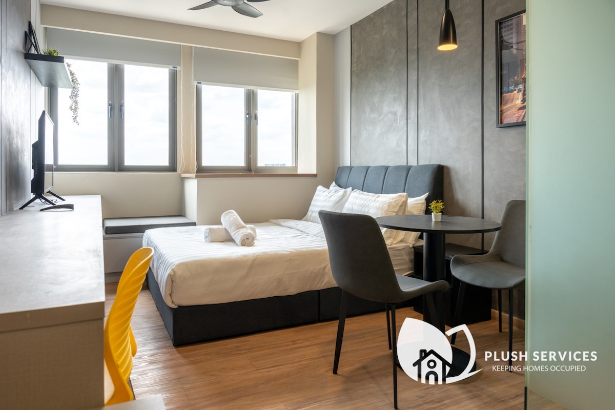 A dual-studio apartment showcases a spacious sleeping area with a queen bed and fresh linens. A sleek desk and dining table are positioned near the windows, which provide ample natural light. A television is mounted on the wall, enhancing the living space's modern design.