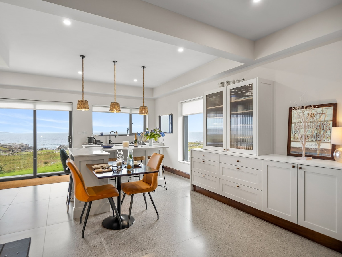 A contemporary dining area is set with a table and chairs, showcasing a serene view of the ocean through large glass doors. Modern pendant lights hang above, while a side cabinet provides additional storage. The space is bright and inviting, with a subtle color palette.