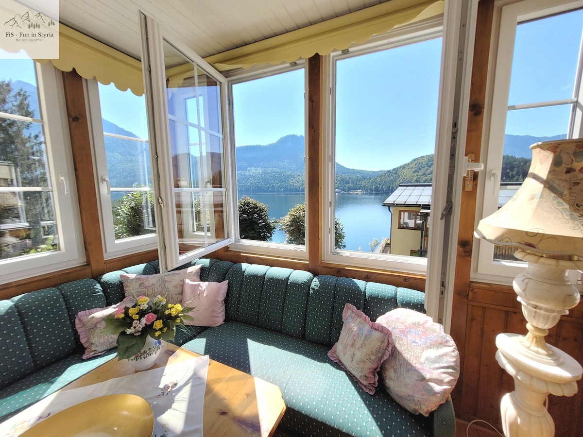 A sunlit glazed veranda showcases panoramic views of the surrounding mountains and Altausseer Lake. A cozy corner sofa is adorned with cushions and a floral arrangement on the table, creating a peaceful setting for relaxation and enjoyment of the natural scenery.