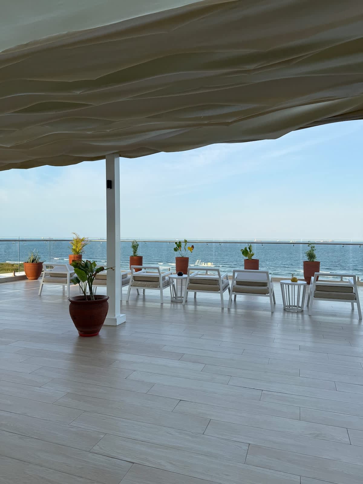 An outdoor terrace features several white lounge chairs arranged alongside potted plants. A calm sea is visible in the background, framed by a clear sky. Light wood flooring extends across the area, creating a spacious and inviting setting.